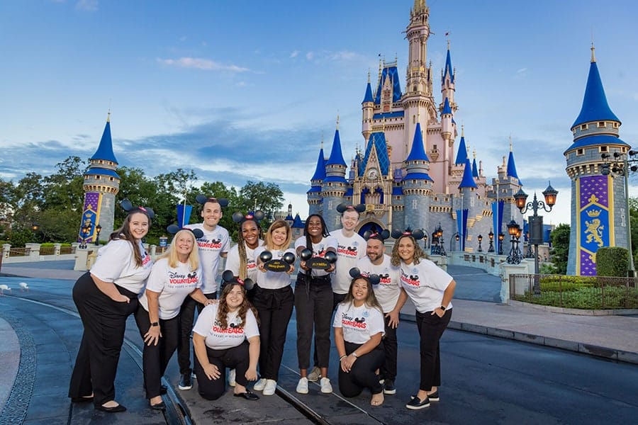 Disney volunteers and cast members pose in front of Cinderella Castle holding Mickey ear hats.
