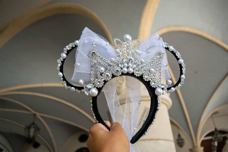 Hand holding the Minnie Mouse Black Velvet ear headband featuring a combination of pronged faceted glass crystals, acrylic crystals, and synthetic pearls hand-stitched onto the headband and ears. A large off-white organza bow and a Silver plated metal tiara adorned with more glass crystals, pearls and beads.