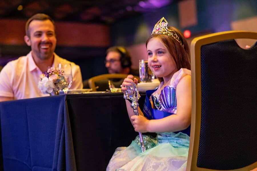 Young female wish kid dressed like Ariel holds a septer while is seated at table with her father looking at her