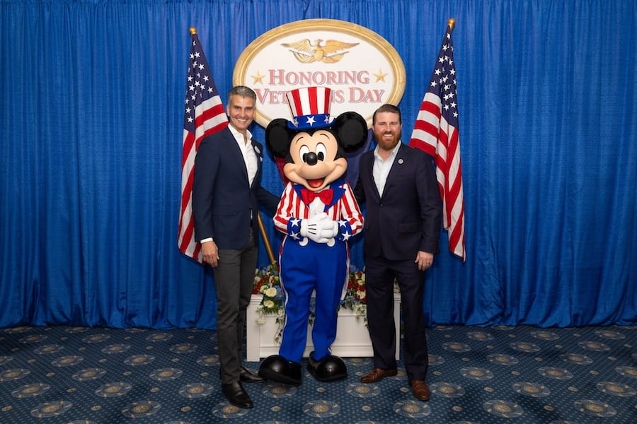 Photo of Josh D’Amaro, Chairman of Disney Experiences with Jared Lyon, president and CEO of Student Veterans of America with Patriotic Mickey Mouse and a sign that says “Honoring Veterans Day”