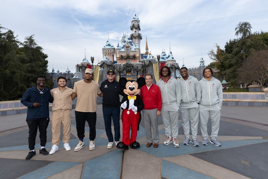 Michigan and Alabama team and coaches commemorative group photo with Mickey Mouse in front of Sleeping Beauty’s Winter Castle at Disneyland