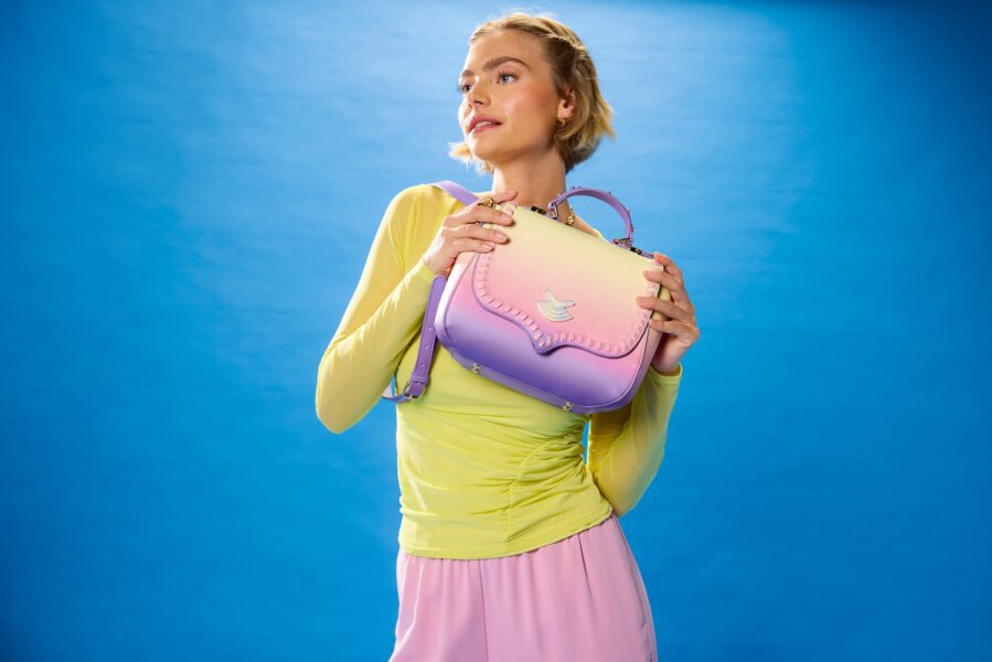 Woman wearing pastel yellow top and pink skirt holds a gradient pink to yellow handbag against a solid blue background posing