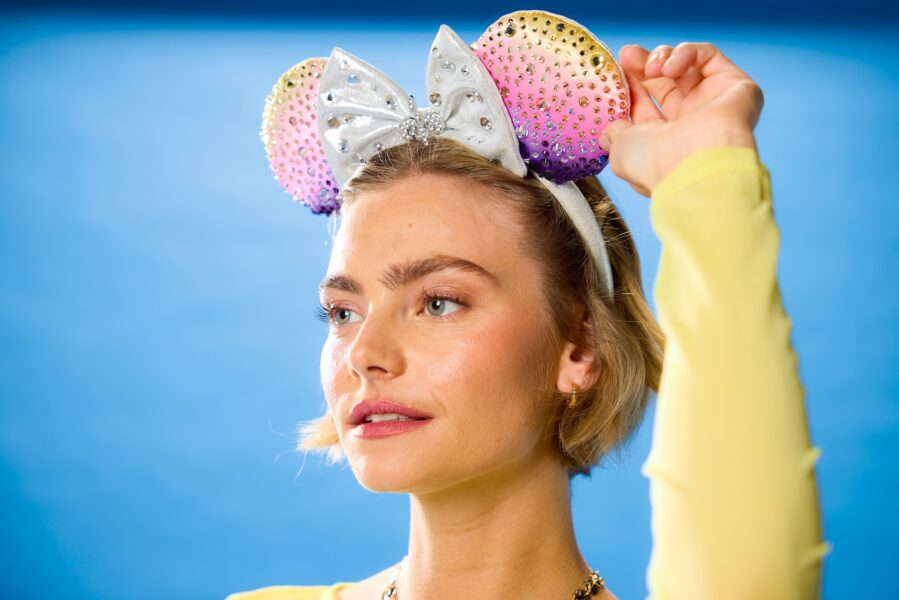 Close up portrait of a woman wearing a rhinestone ears headband with a silver bow being adjusted by a hand against a blue background