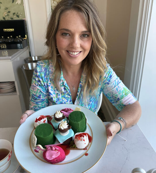 Smiling woman selfie with a plate of colorful cupcakes and desserts in front of her in a bright kitchen green cakes and pink frosting visible
