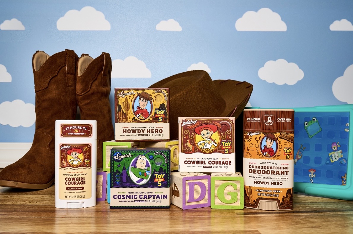 Cowboy Boots and Assorted Toy Story themed Soap Boxes Arranged on a Wooden Surface with Alphabet Blocks Spelling Dg Against a Blue Cloud wall Backdrop