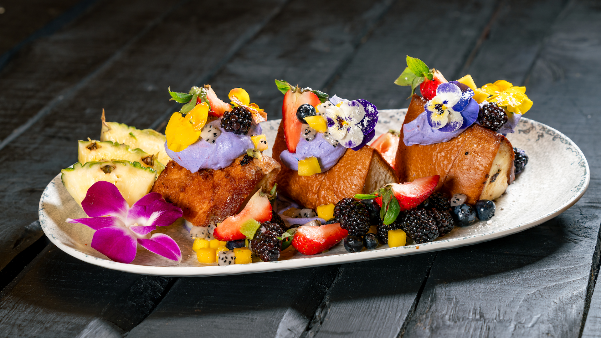 2026DLRTraderSams15thAnniversary24 | The Main Street Mouse Three Br2026 dlr trader sams 15th anniversary 24ioche French Toast Slices Topped with Purple Cream Fresh Berries Mango Pineapple and Edible Flowers on a White Plate
