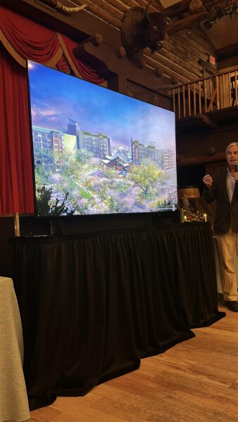 Presenter speaks beside a large screen displaying a modern architectural rendering in a rustic lodge like venue with red curtains and a mounted elk head above