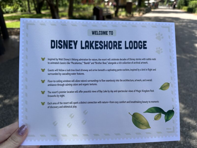 Sign reading Welcome to Disney Lakeshore Lodge with decorative border held outdoors in a park setting
