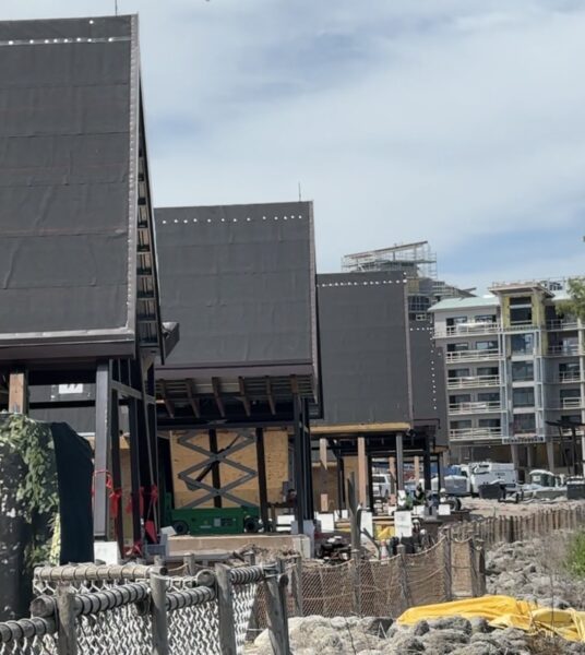 Row of dark modular building units on steel supports under construction along a body of water with a chain link fence and construction debris in the foreground and residential buildings in the background