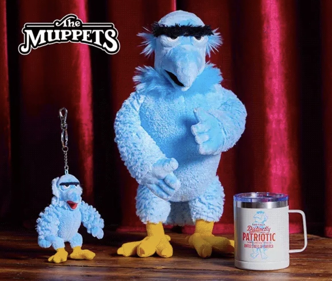 Two Blue Muppet Characters a Large One Giving a Thumbs Up and a Small Keychain Version on a Wooden Surface with Red Curtains and a Patriotic Mug in View
