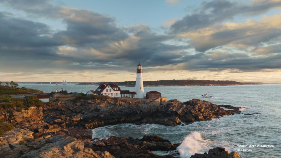 IMG1592 | The Main Street Mouse Lighthouse on a Rocky Coastline with White Building Adjacent Calm Sea and Sailboats in the Distance at Sunset © Disney