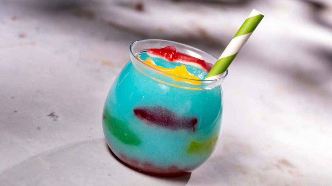 Blue Tropical Drink in a Curved Glass with a Green and white Striped Straw and Red yellow Garnish on a Light Surface