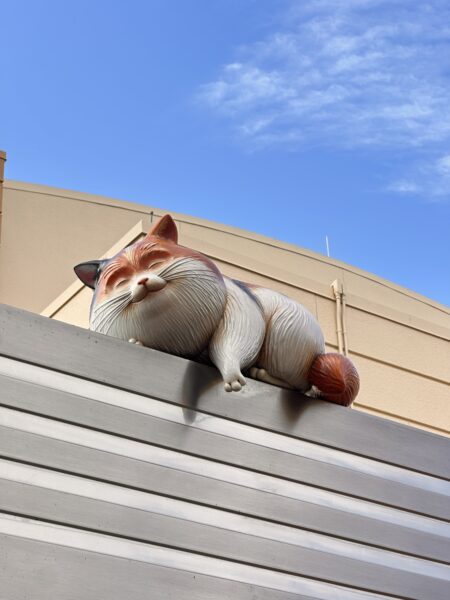Large Orange and white Cat Statue Lying Curled on a White Railing with a Blue Sky Behind It Outdoors