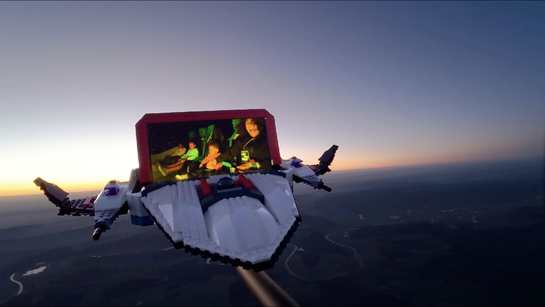 LEGO spaceship with a red framed screen showing smiling people flying over a coastline at sunset