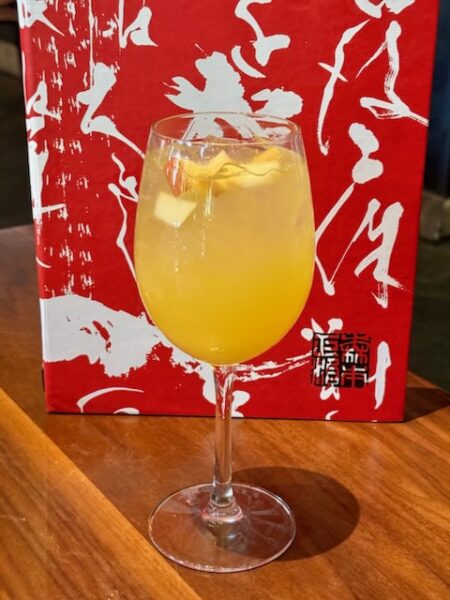 Orange Cocktail with Ice in a Stemmed Glass Lemon Twist Garnish on a Wooden Table with a Red Abstract Backdrop