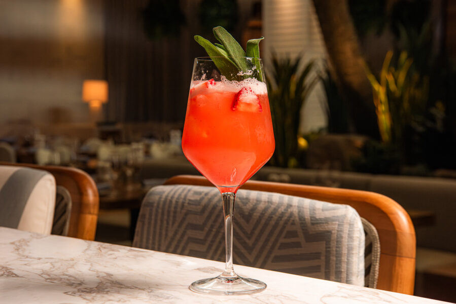 Pink Cocktail in a Stemmed Glass with Ice and a Green Herb Garnish on a Marble Table Restaurant Setting in the Background