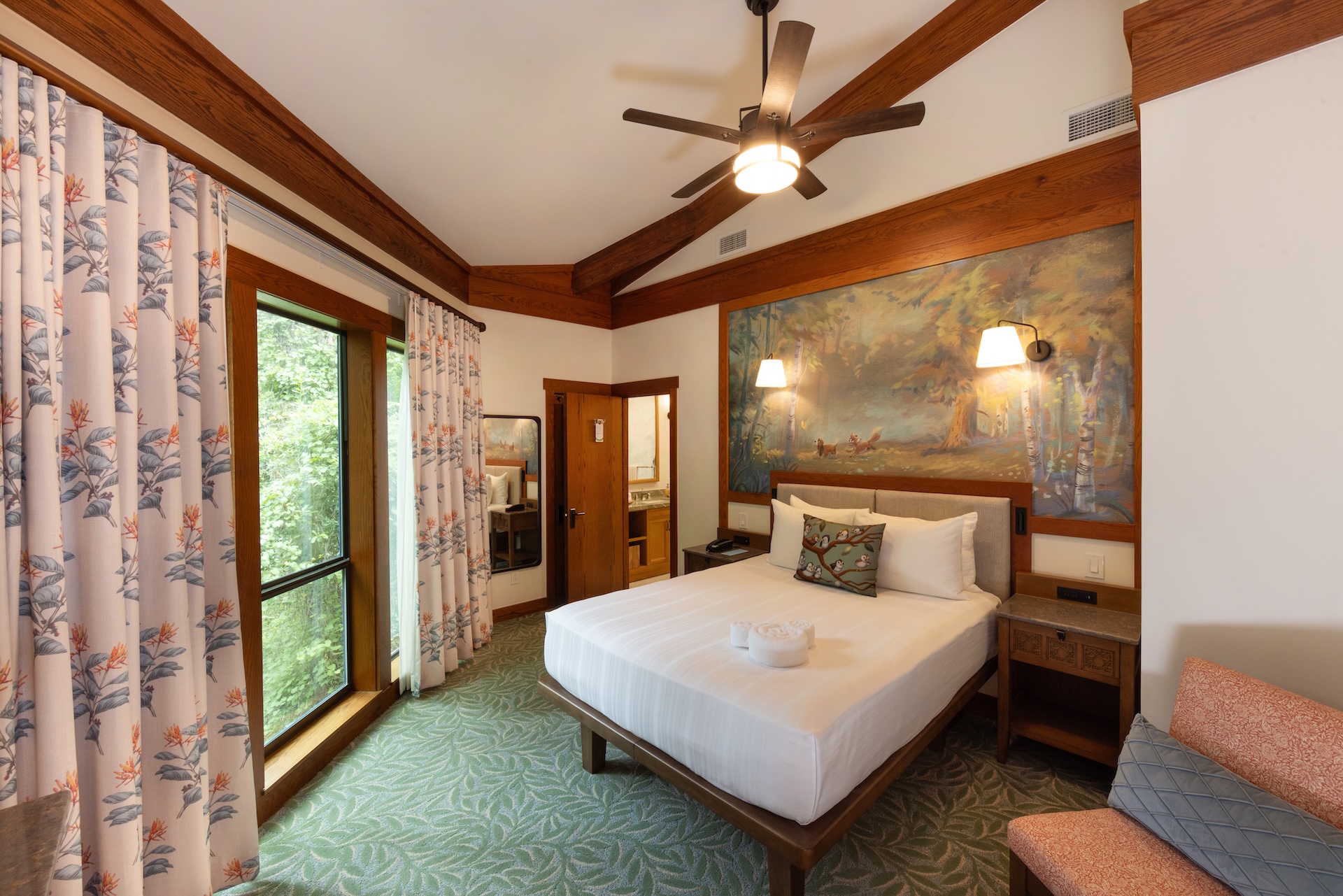 2026DVCReimaginedTreehouseVillas10 | The Main Street Mouse Hotel Room Featuring a King Bed with White Linens Wooden Headboard and a Large Forest Mural Above the Bed Window with Floral Curtains to the Left and Ceiling Fan Overhead