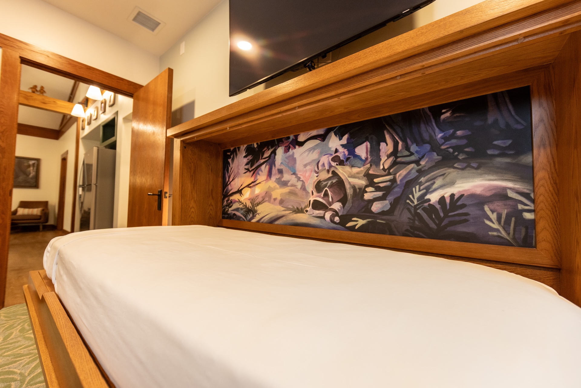 2026DVCReimaginedTreehouseVillas12 | The Main Street Mouse Low angle View of a Large Bed with a Wooden Headboard and a Colorful Mural Panel Behind It Tv Mounted Above Doorway and Hallway to the Left
