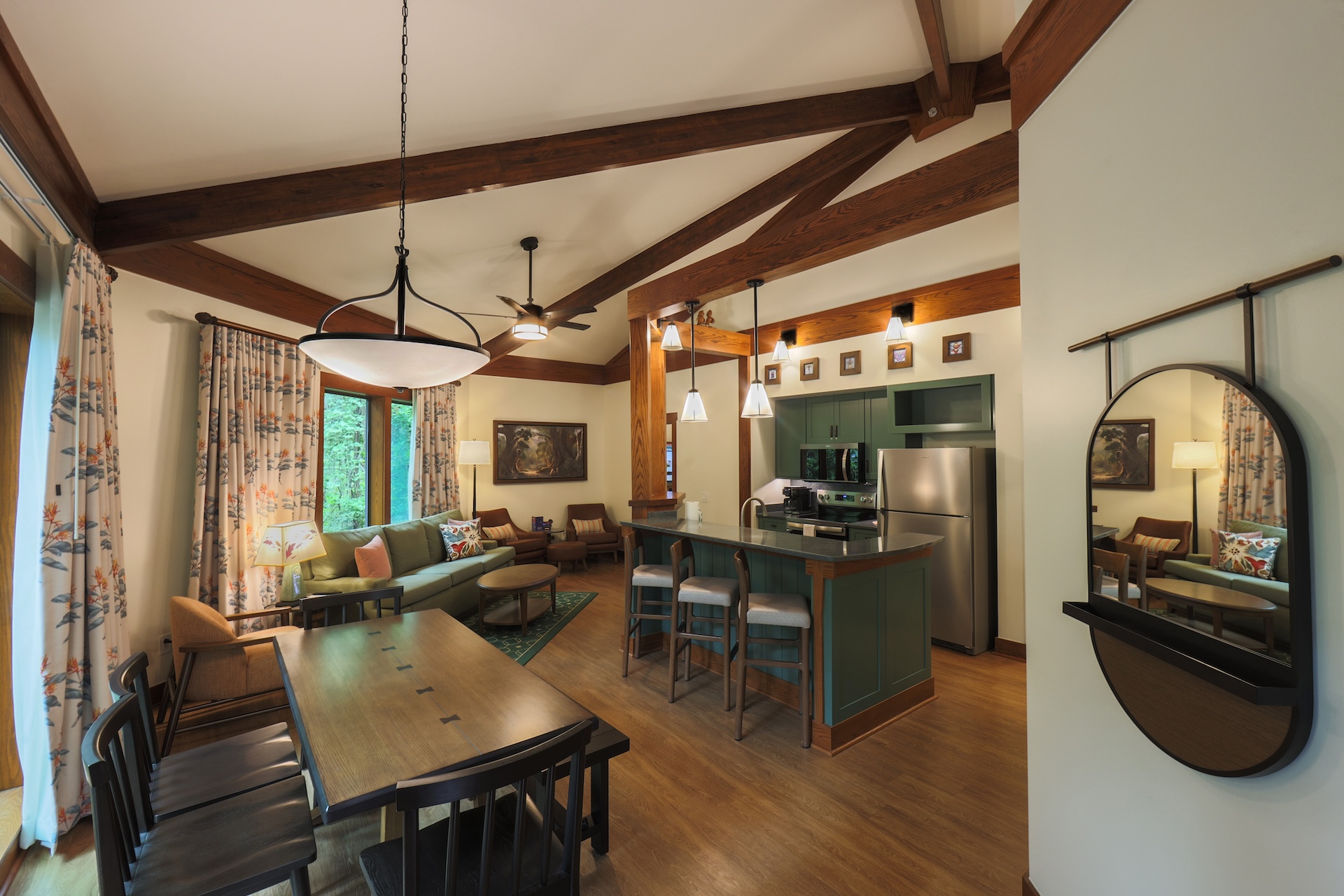 2026DVCReimaginedTreehouseVillas15 | The Main Street Mouse Open plan Living Area with Exposed Wooden Beams a Green Kitchen Island and a Dining Table Large Windows with Floral Curtains on the Left and a Round Wall Mirror on the Right