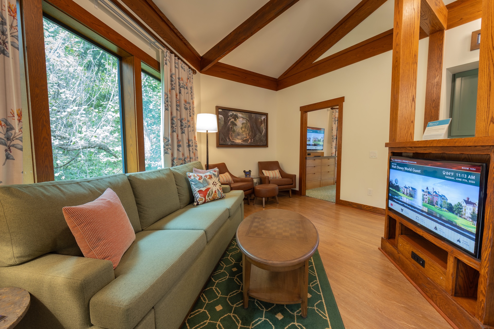 2026DVCReimaginedTreehouseVillas16 | The Main Street Mouse Living Room with a Green Sectional Sofa Pink Pillows and a Round Wooden Coffee Table Near Large Windows Showing Green Trees Outside
