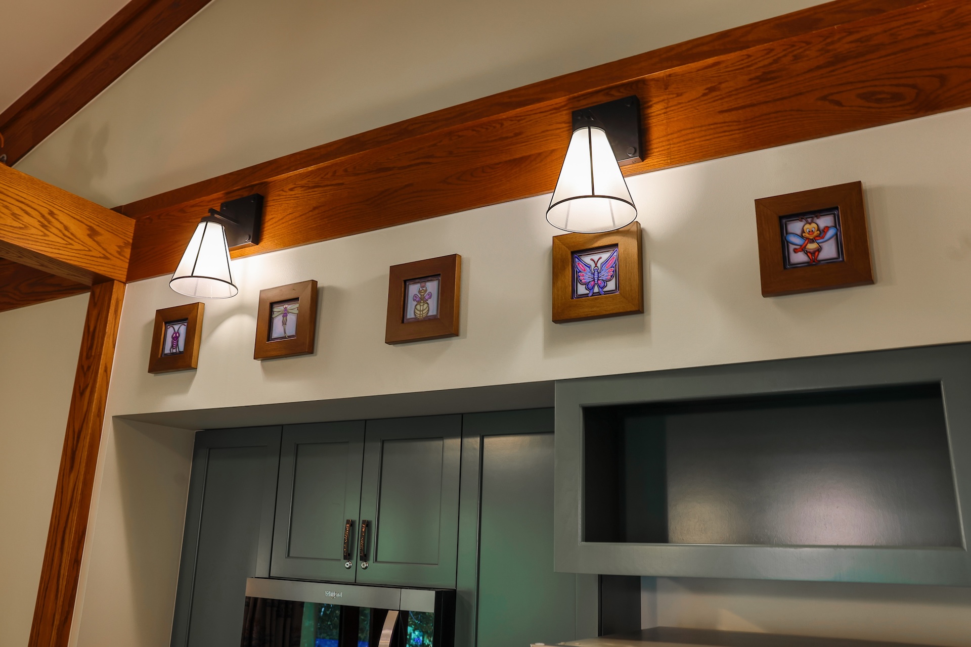 2026DVCReimaginedTreehouseVillas4 | The Main Street Mouse Kitchen Area with Gray green Cabinets Wooden Beam and Two Cone shaped Wall Lights Small Wood framed Insect Pictures Line the Wall Above the Cabinets