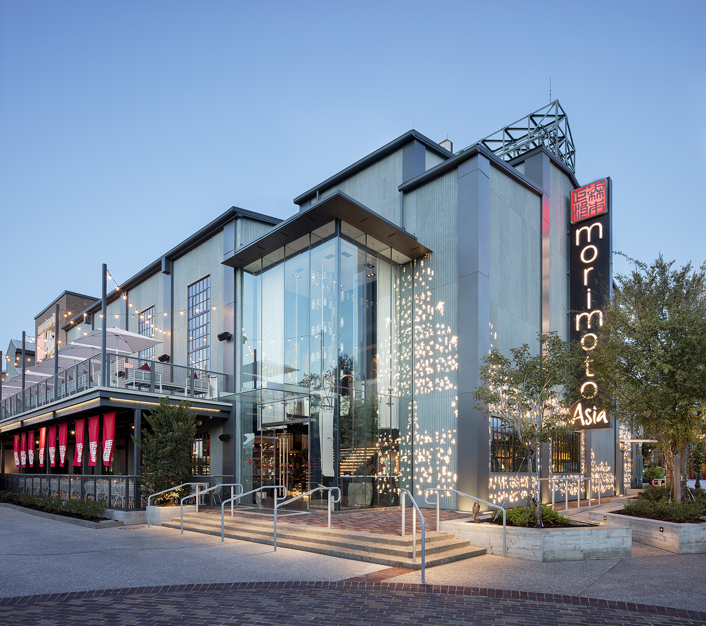 Exterior View of Morimoto Asia Restaurant with a Tall Vertical Sign Glass Atrium and String Lights at Dusk