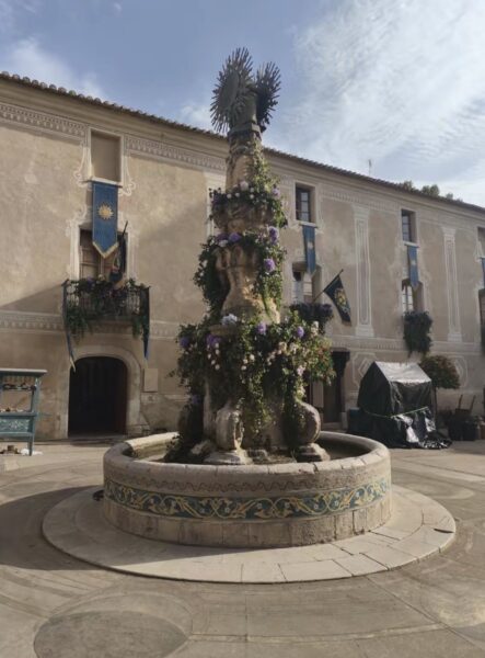 IMG0007 | The Main Street Mouse Decorative Stone Fountain in a Circular Basin Covered with Flowers in a Historic Town Square with a Pale Building and Blue Banners