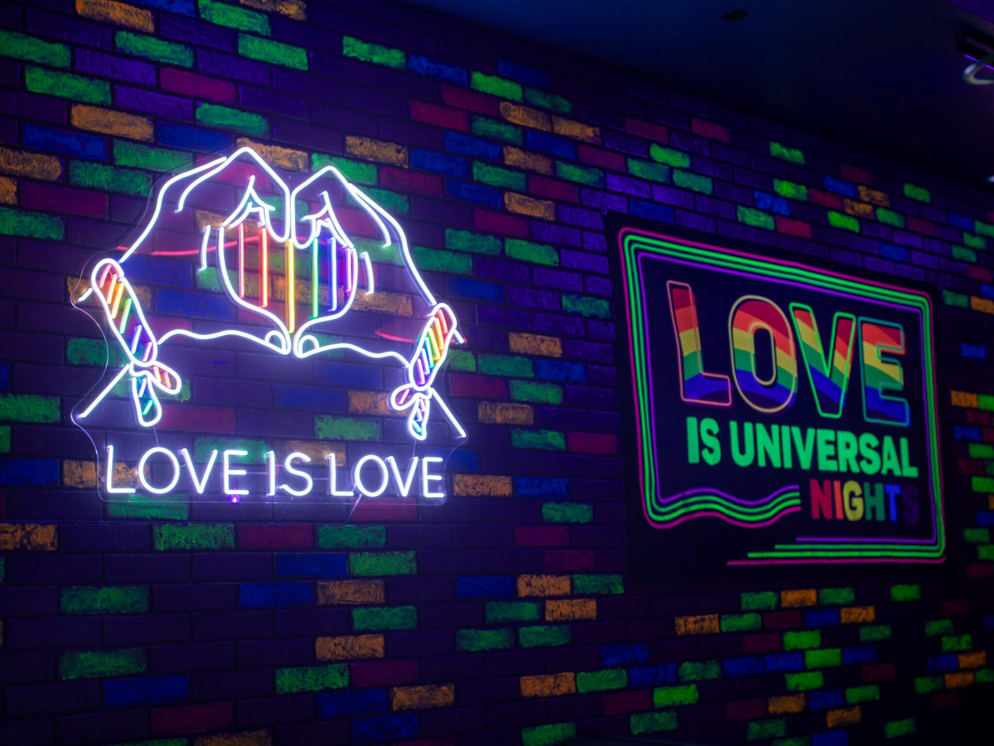 LoveisUniversalSign22048x1536 | The Main Street Mouse Neon Sign with White Outlined Hands and the Words love is Love Against a Colorful Brick Wall a Second Neon Sign Reads love is Universal in Bright Colors to the Right