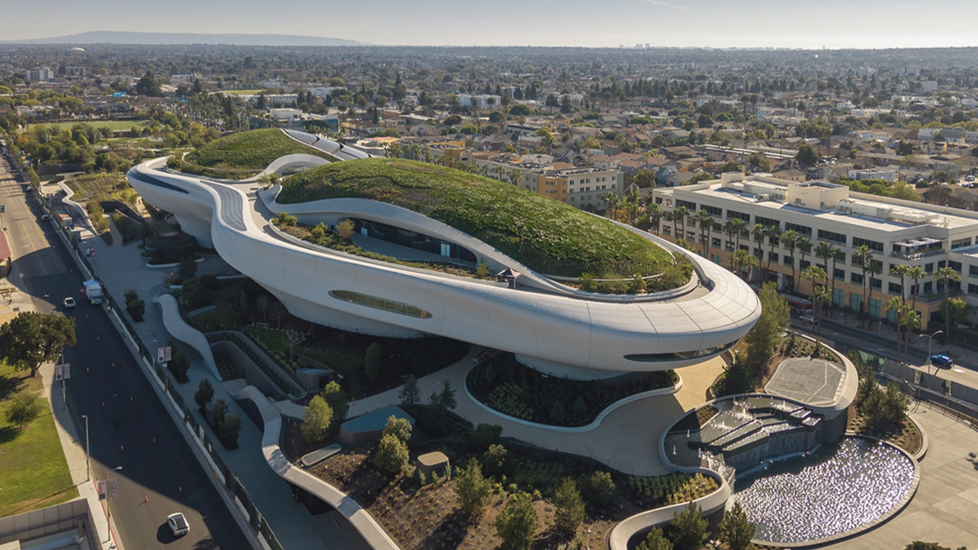 Aerial View of a Futuristic White Building with Curving Green Roofs and Spiral Walkways Set in an Urban Landscape